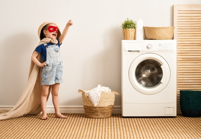 Little boy with superhero mask and blanket cape next to washing machine.