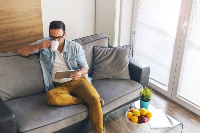 Man sitting on couch looking at tablet while drinking coffee.