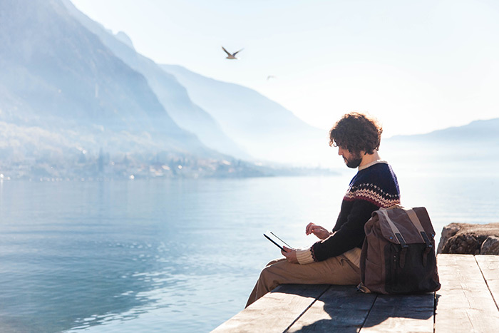 Man working on laptop while sitting on edge of dock by the lake.