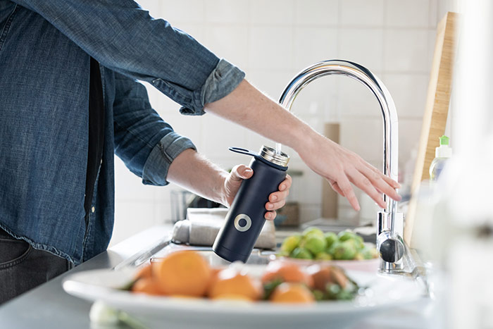 Person filling water bottle at sink with fruit on the counter.