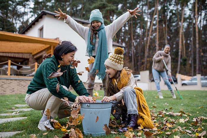 Grandmother, mother and two young daughters raking and playing in leaves outside.