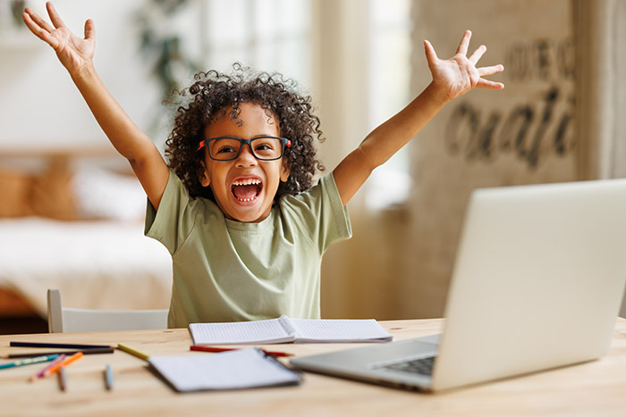 Excited little boy smiling with arms raised doing homework at table with laptop.