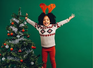 Girl standing next to Christmas tree wearing red reindeer antlers and wearing Christmas sweater.