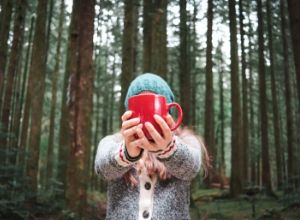 Woman standing in woods holding red coffee mug.