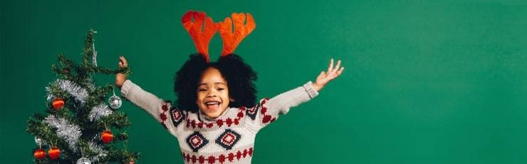 Young girl standing next to Christmas tree wearing Christmas sweater and reindeer antlers.